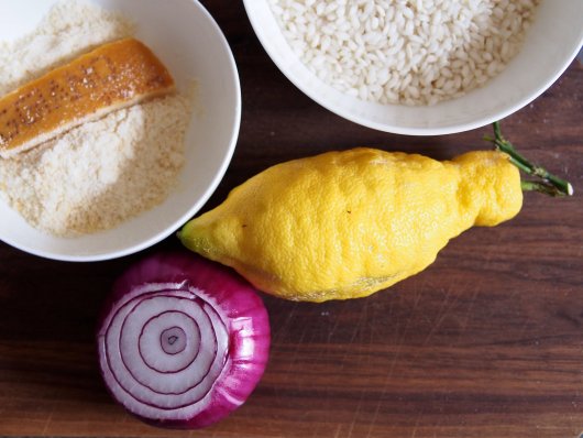 Zutaten für Risotto ai gamberi e cedri Eine Schüssel mit geriebenem Parmesan, eine Schüssel mit Risotto-Reis, eine rote Zwiebel aus Tropea und eine Cedro auf einem Holzbrett