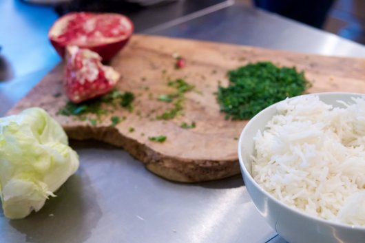Eine Schüssel Reis und ein Salatkopf, im Hintergrund ein Schneidbrett mit einem halbierten Granatapfel und gehacktem Koriander.
