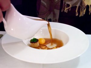 Essenz vom Almo, Dotter vom Herk-Ei, Knödel, Sherry II Consommé wird in einen Teller mit Einlagen eingeschenkt.